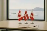 Флаги Канады, Дании и Гренландии на столе переговоров у окна с видом на арктическое побережье.