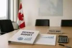 Папка «Defence Budget» с графиками на столе в офисе, на фоне размыты флаг Канады и карта Арктики