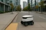 Небольшой робот-доставщик стоит на тротуаре Ванкувера у тактильной плитки, мягкий дневной свет.