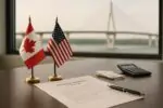 Документ о сделке и маленькие флаги Канады и США на столе, на фоне размытый мост через реку.