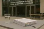 Стакан чистой воды и папка с документами на фоне современного зала встреч в Оттаве, без людей