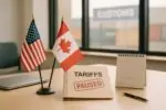 Флаги США и Канады на столе рядом с документами и штампом «пауза», на фоне размытая логистика