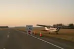 Небольшая красно-белая Cessna стоит на обочине сельской трассы на рассвете, рядом конусы и знак.