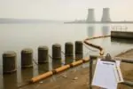 Берег озера с водозаборными решётками и несколькими рыбами; вдали видна АЭС в лёгкой дымке.