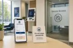 Светлый интерьер банка: смартфон с экраном входа в онлайн-банк и табличка «не делитесь паролем», символ ограничений для ИИ.